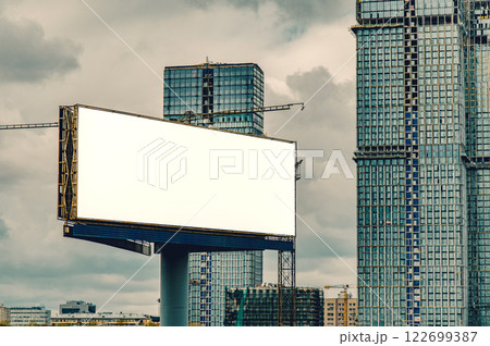 A modern urban landscape with a blank white billboard set against the backdrop of under-construction skyscrapers 122699387