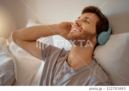 Man listening to music, podcast or radio with headphones at home while relaxing, smile and happy in his bedroom. Peaceful, freedom and young male streaming an online audio playlist enjoying weekend 122701663