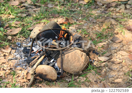 A classic three legged iron stand is placed over an open fire, designed to hold cooking pots. This method reflects traditional outdoor cooking, commonly used in camping and rural kitchens. A classic three legged iron stand is placed over an open fire, designed to hold cooking pots. This method reflects traditional outdoor cooking, commonly used in camping and rural kitchens. 122702589