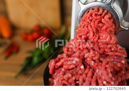 Concept of healthy and proper nutrition, cooking.Close-up of red ground beef with fat streaks coming out of a metal meat grinder on a wooden background, vegetables and herbs are visible out of focus Concept of healthy and proper nutrition, cooking.Close-up of red ground beef with fat streaks coming out of a metal meat grinder on a wooden background, vegetables and herbs are visible out of focus 122702643