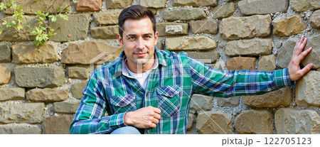 Young man poses casually against a rustic stone wall in a peaceful outdoor setting during daylight 122705123