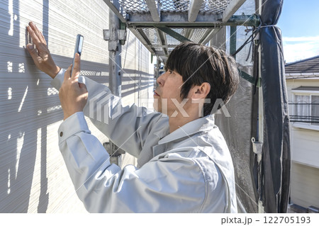 戸建て家屋の外壁重ね張りリフォーム工事　携帯で記録する男性現場監督者 122705193