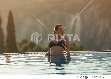 Slim woman in swimsuit walking by the edge of luxury infinity pool with jungle view  122705705