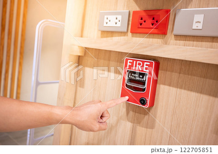 Hand pointing at red fire alarm switch on wooden wall in the office interior with electric sockets 122705851