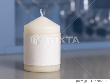 Cream pillar candle with a label on grey table against blue wine glasses holder closeup, mockup Cream pillar candle with a label on grey table against blue wine glasses holder closeup, mockup 122706302
