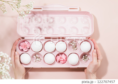 Female hands holding pink egg carton filled with white eggs and delicate pink flowers on pastel pink background. Soft and creative design perfect for holiday Easter and spring-inspired themes Female hands holding pink egg carton filled with white eggs and delicate pink flowers on pastel pink background. Soft and creative design perfect for holiday Easter and spring-inspired themes 122707457