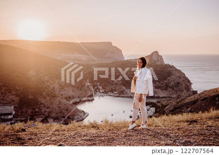 A woman stands on a hill overlooking a body of water. The sun is setting in the background, casting a warm glow over the scene. The woman is enjoying the view and taking in the beauty of the landscape A woman stands on a hill overlooking a body of water. The sun is setting in the background, casting a warm glow over the scene. The woman is enjoying the view and taking in the beauty of the landscape 122707564