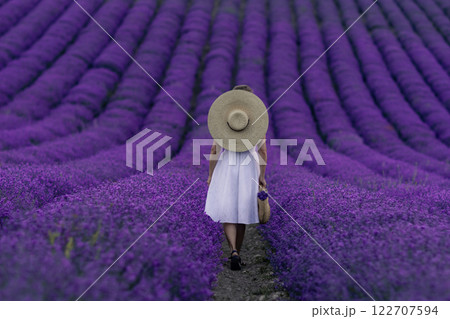 A woman wearing a straw hat walks through a field of purple flowers 122707594