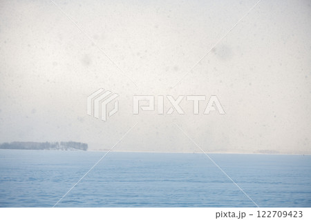 Snow is gently falling on a vast frozen lake on a cold winter day, creating a serene and peaceful atmosphere with a distant island covered in trees Snow is gently falling on a vast frozen lake on a cold winter day, creating a serene and peaceful atmosphere with a distant island covered in trees 122709423