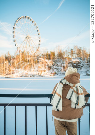 Cute girl dressed in cozy winter attire, including a beanie and scarf, gazing at a colorful ferris wheel beyond a frozen lake on a bright, sunny winter day Cute girl dressed in cozy winter attire, including a beanie and scarf, gazing at a colorful ferris wheel beyond a frozen lake on a bright, sunny winter day 122709521