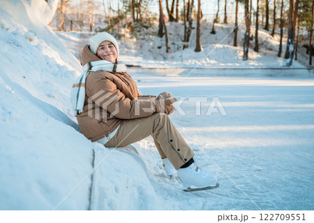 cute girl wearing winter clothes sitting on snowbank by frozen lake, resting after ice skating in beautiful winter forest, enjoying winter sports and outdoor activities on sunny day cute girl wearing winter clothes sitting on snowbank by frozen lake, resting after ice skating in beautiful winter forest, enjoying winter sports and outdoor activities on sunny day 122709551