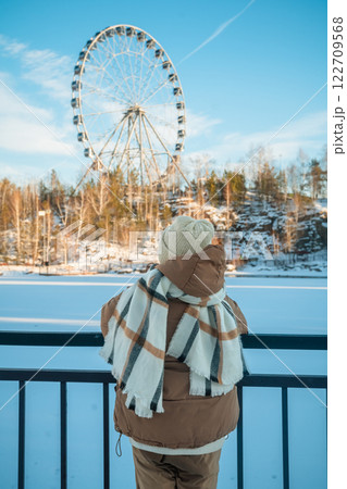 Cute girl dressed in cozy winter attire, including a beanie and scarf, gazing at a colorful ferris wheel beyond a frozen lake on a bright, sunny winter day 122709568