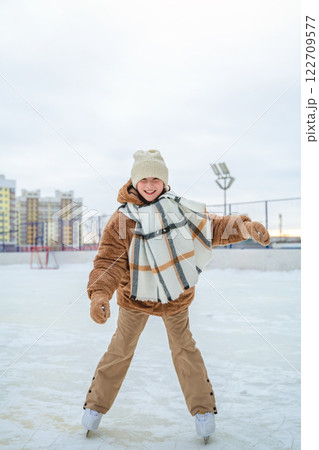 Smiling girl on skates, dressed in warm winter clothes, gracefully glides on the outdoor skating rink, enjoying the joy of the winter season and fun activities in the snow. Smiling girl on skates, dressed in warm winter clothes, gracefully glides on the outdoor skating rink, enjoying the joy of the winter season and fun activities in the snow. 122709577