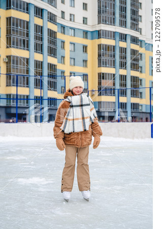 Girl wearing warm winter clothes ice skating on a rink in front of a residential building, enjoying the thrill of winter sports and embracing a fun outdoor leisure activity Girl wearing warm winter clothes ice skating on a rink in front of a residential building, enjoying the thrill of winter sports and embracing a fun outdoor leisure activity 122709578