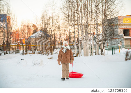 Warmly dressed girl pulling an inflatable snow tube while walking along a snowy path in a winter park, eagerly preparing for a day filled with fun sledding and winter activities 122709593