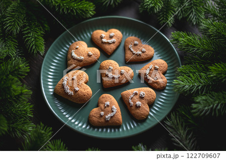 Heart-shaped gingerbread cookies with smiley faces made of icing sugar pearls rest on a green plate surrounded by fir branches, creating a festive christmas scene Heart-shaped gingerbread cookies with smiley faces made of icing sugar pearls rest on a green plate surrounded by fir branches, creating a festive christmas scene 122709607