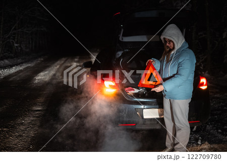 On a snowy night road, a woman sets a warning triangle after her car breaks down in a winter forest. Her car's trunk is open, and steam rises from the cold, creating a dramatic scene. 122709780