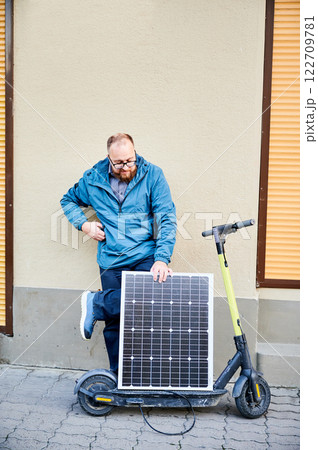 Male person holds photovoltaic solar panel, for charging electric scooter in urban settings. Integration of solar power as sustainable energy source, promoting eco-friendly urban transportation. Male person holds photovoltaic solar panel, for charging electric scooter in urban settings. Integration of solar power as sustainable energy source, promoting eco-friendly urban transportation. 122709781