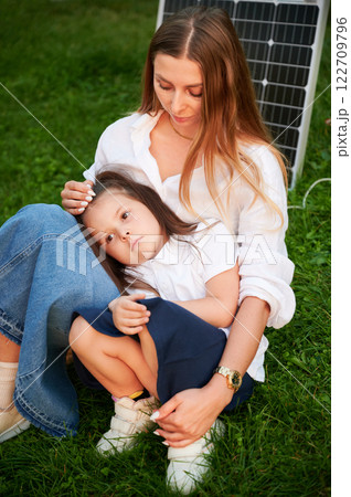 Mother and daughter sit together on grass, embracing in warm moment with solar panel beside in the evening. Concept of renewable energy and family, highlighting commitment to sustainable future. Mother and daughter sit together on grass, embracing in warm moment with solar panel beside in the evening. Concept of renewable energy and family, highlighting commitment to sustainable future. 122709796