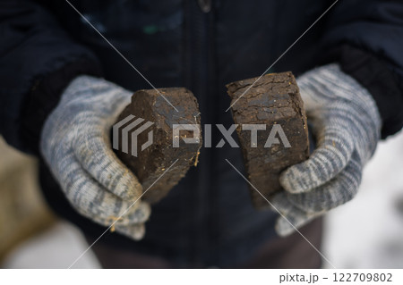 A large plan of peat briquettes for kindling a stove in hands. A farmer in gloves holds two peat briquettes for kindling a stove in winter, which is a sustainable and traditional heating solution. A large plan of peat briquettes for kindling a stove in hands. A farmer in gloves holds two peat briquettes for kindling a stove in winter, which is a sustainable and traditional heating solution. 122709802