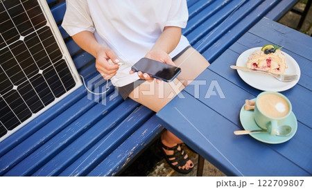 Cropped photo of girl putting phone on charge from solar battery. Young girl relaxing in cafe while charging smartphone from solar battery charger. Concept of alternative energy and green lifestyle. 122709807