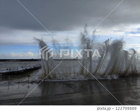 Waves crash against the shore creating splashes near a coastal walkway during a cloudy day Waves crash against the shore creating splashes near a coastal walkway during a cloudy day 122709889