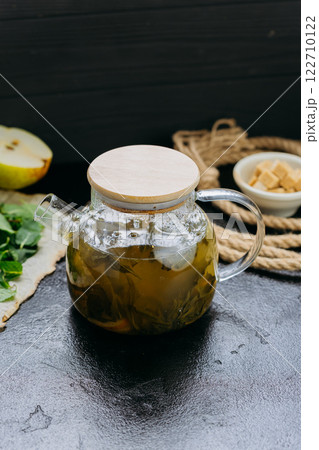 Pear and mint herbal tea in cup with glass teapot, fresh ingredients, and sugar cubes on dark table 122710122
