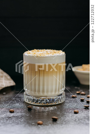 Layered latte with frothy milk and crushed nuts in crystal glass with coffee beans on dark table 122712032