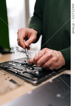 Technician male repairing laptop motherboard with screwdriver, surrounded by various components and tools on workbench, close-up. Concept of computer hardware, repairing, upgrade and technology. 122712304