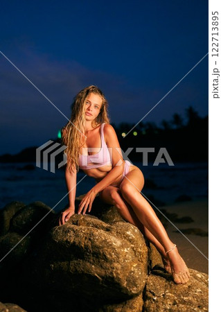 Stunning woman model in bikini at night beach. Ocean waves crash in dark background. Perfect fit body, relaxing, posing on large rocks. Fashion, beauty, fitness, travel and exotic vacation theme. 122713895