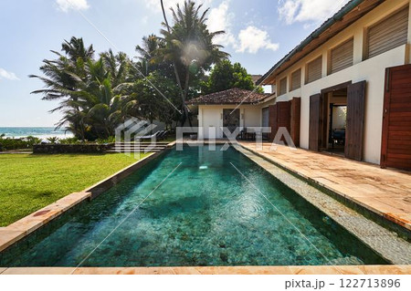 Sunny tropical villa with pool, palm trees by ocean. Vintage architecture, wooden shutters open. Relaxing holiday vibe in exotic paradise. Poolside chairs invite leisure. Charming getaway spot. Sunny tropical villa with pool, palm trees by ocean. Vintage architecture, wooden shutters open. Relaxing holiday vibe in exotic paradise. Poolside chairs invite leisure. Charming getaway spot. 122713896