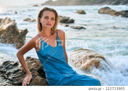 Model woman sits on cliff in wet dress at exotic island ocean. Stunning femininity, divine beauty with storming waves. Perfect for travel, nature, fashion themes. Female model, elegance by sea. 122713905