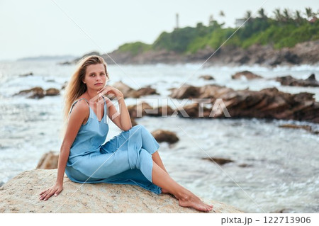 Blonde woman sits on rocky beach, ocean view. Wearing blue dress, relaxed. Waves crash on rocks. Tropical coastline in background, cloudy sky. Ideal for luxury travel fashion. Exotic, serene setting. 122713906