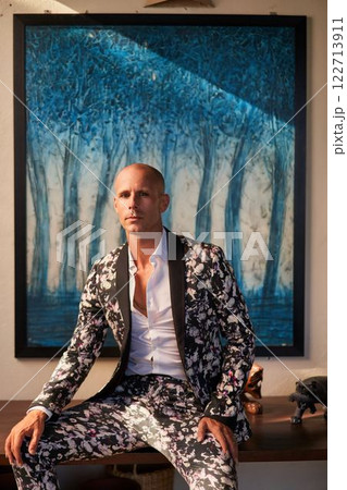 Bald man in bold floral suit sits at villa, possible groom outfit. Elegant gay fashion, unique style for weddings. Trendy chic look, modern expression. Luxury setting indoors, artistic blue painting. Bald man in bold floral suit sits at villa, possible groom outfit. Elegant gay fashion, unique style for weddings. Trendy chic look, modern expression. Luxury setting indoors, artistic blue painting. 122713911