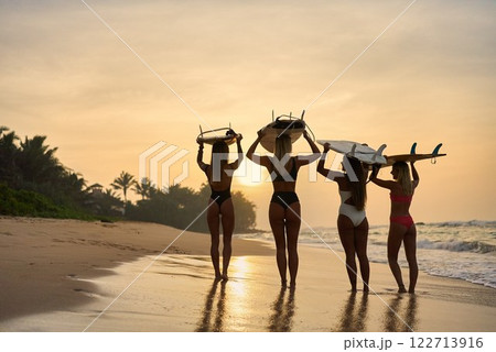 Group of women carry surfboards walking on sandy beach. Sunset over ocean waves. Friends enjoy surf adventure. Surf camp travel. Beach lifestyle holiday. 122713916