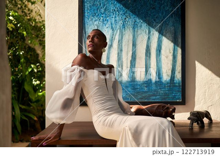 Person in elegant white dress sits in villa. Gender fluid black bride enjoys sunny day indoors. LGBTQ wedding themes, inclusive love. Perfect image for gay-friendly hotels, event agencies. Person in elegant white dress sits in villa. Gender fluid black bride enjoys sunny day indoors. LGBTQ wedding themes, inclusive love. Perfect image for gay-friendly hotels, event agencies. 122713919