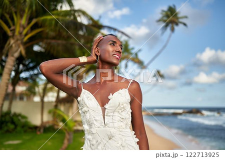 Gender fluid person in elegant wedding dress, serene tropical beach. Ocean waves, palm trees swaying. LGBTQ-friendly bridal moment. Inclusive destination ceremony. Unique non-binary bridal fashion. 122713925