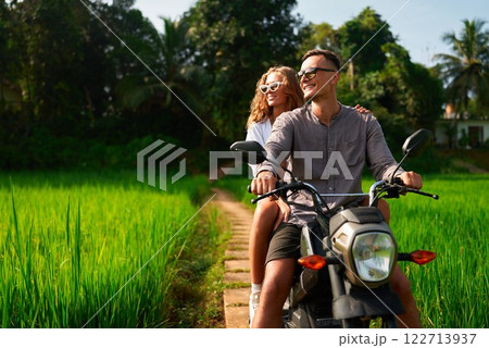 Couple rides motorbike on rural path by rich rice fields. Man drives scooter with woman enjoying green scenery. Romantic adventure amid nature. Explore countryside, feel freedom. Travel concept. Couple rides motorbike on rural path by rich rice fields. Man drives scooter with woman enjoying green scenery. Romantic adventure amid nature. Explore countryside, feel freedom. Travel concept. 122713937