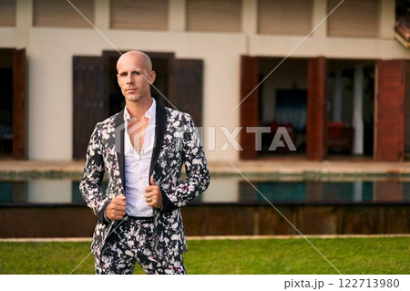 Bald man, bold floral suit poses villa courtyard. Stylish outfit, modern groom trends. Luxury poolside. Elegant attire, fashion statement. Perfect for weddings, upscale events, fashion, LGBTQ. Bald man, bold floral suit poses villa courtyard. Stylish outfit, modern groom trends. Luxury poolside. Elegant attire, fashion statement. Perfect for weddings, upscale events, fashion, LGBTQ. 122713980