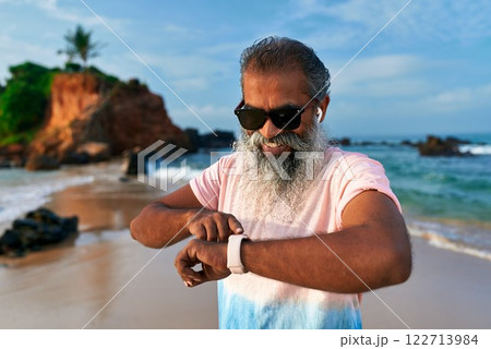 Gray bearded senior man with sunglasses uses smart watch on beach. Mature male with earbuds checks fitness tracker for health. Wireless tech use by elderly for wellbeing. 122713984