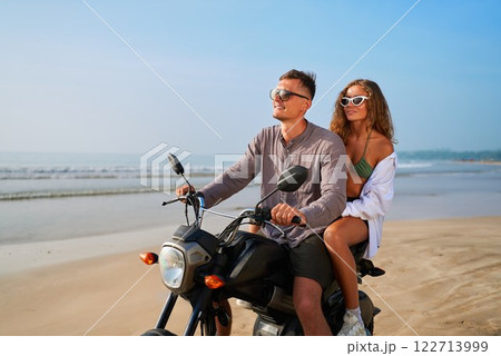 Young couple rides motorbike on beach. Man drives motorcycle with woman sitting behind. Relaxed travel mood. Perfect vacation at ocean shore. Adventure seekers enjoy sun, sea breeze. 122713999
