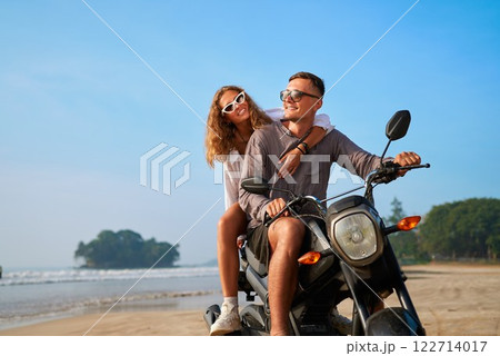 Couple rides motorbike on sunny beach. Girl hugs boyfriend on motorcycle. Happy couple enjoys seaside adventure exploring tropical island. Man drives bike, woman loves ocean breeze. Couple rides motorbike on sunny beach. Girl hugs boyfriend on motorcycle. Happy couple enjoys seaside adventure exploring tropical island. Man drives bike, woman loves ocean breeze. 122714017
