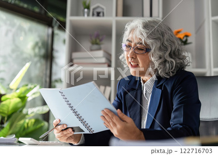 Elderly female business executive analyzing reports and making notes in modern office with green plants 122716703