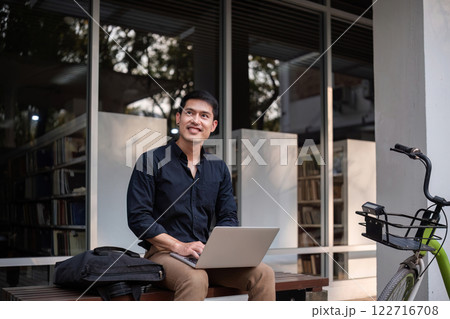 A young Asian businessman rides a bicycle to work. Sitting outside the office working using a laptop. 122716708