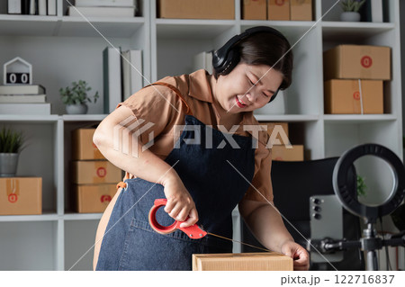 Woman in denim apron using scissors to cut packing tape in a modern workspace, preparing boxes for shipment 122716837