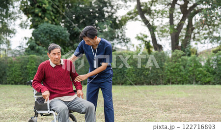 Nursing assistant providing support to elderly man in a wheelchair, emphasizing care in outdoor setting 122716893