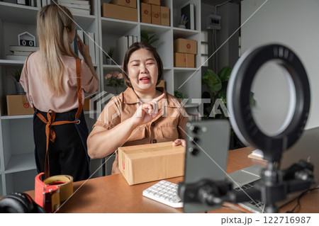 E-commerce entrepreneur explaining shipment process on camera with package, while assistant organizes inventory E-commerce entrepreneur explaining shipment process on camera with package, while assistant organizes inventory 122716987