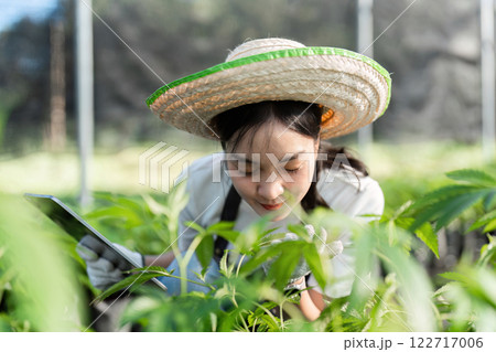 Young Female Farmer Examining Plants in a Greenhouse with a Tablet and Straw Hat 122717006
