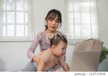 Focused mother assisting her baby while working on a laptop at home. Focused mother assisting her baby while working on a laptop at home. 122717049