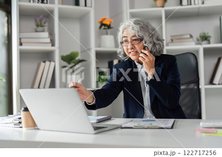 Elderly business woman effectively communicating during a phone call while working on her laptop in a modern office 122717256